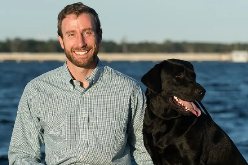Dr. MacKenzie Farnham sitting beside his black labrador, Nugget, by the water.